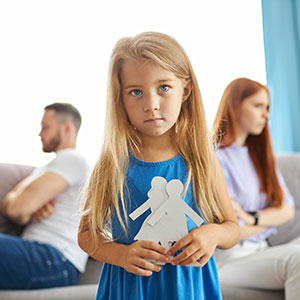 A young girl holding a paper cutout of a family, her parents sitting apart in the back, represent a child custody case.