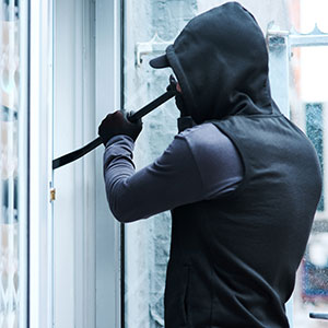 A person in a hoodie using a crowbar to break into a door, representing burglary and theft.