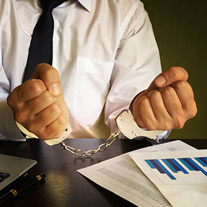 Man in handcuffs at a desk with legal documents, symbolizing white-collar crimes & fraud defense.